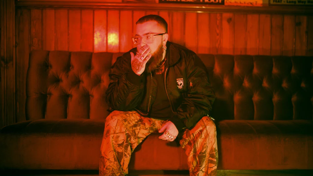 Photo of Riot Code leaning sitting on an old sofa in a pub, leaning forward with the lower half of his face in his hand