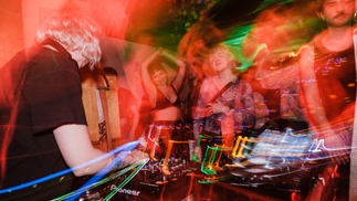 Photo taken from behind the decks at Ar Ais Arís, where numerous people are dancing amid a red motion blur