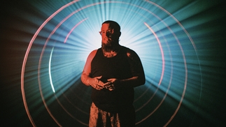 Photo of RIOT CODE wearing a blank tank top in a dark room, with a large beam of light shining around him from the back