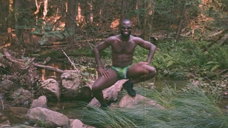 Akanbi crouching on a rock in a river, surrounded by lush woodland. He's wearing black trainers, shades and green speedos