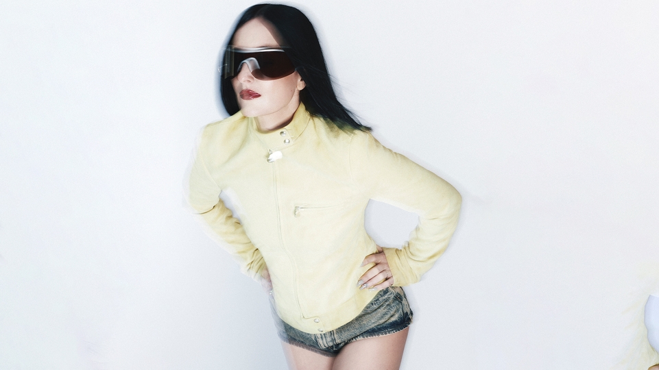 Photo of Anetha wearing a yellow parka and black wrap-around sunglasses against a white background